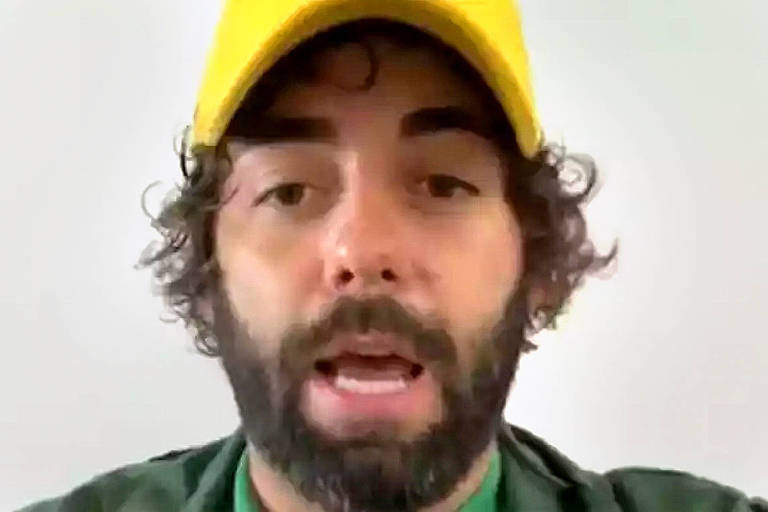 A foto mostra o empresário bolsonarista Esdras Jonatas dos Santos, investigado por suspeita de incentivar agressões a repórteres em Belo Horizonte. Ele usa barba e está de boné amarelo e blusa verde.