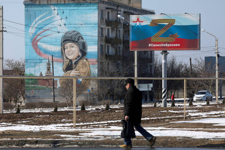 Pedestre caminha perto de outdoor com letra Z, que simboliza a defesa da vitória da Rússia na guerra, e mural com ilustração de criança, na Crimeia 