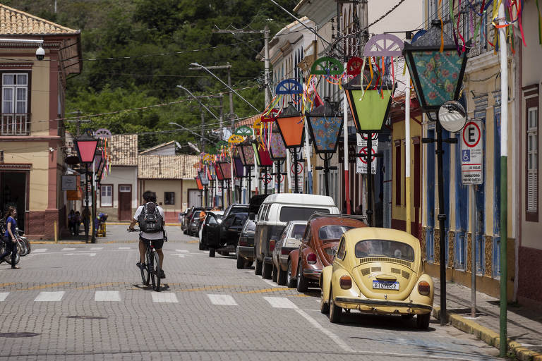 foto mostra rua da cidade com casas antigas, bem pintadas, e fitas e bandeiras nos postes, decoração de carnaval. veículos estacionados e uma pessoa andando de bicicleta, de costas, ao fundo. 