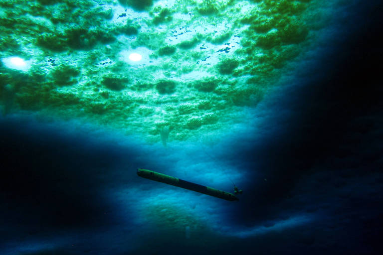 Sombra de um robô estreito e comprido 'nadando' em águas de cor azul escura, logo abaixo de uma 'mancha' verde clara, que é o gelo