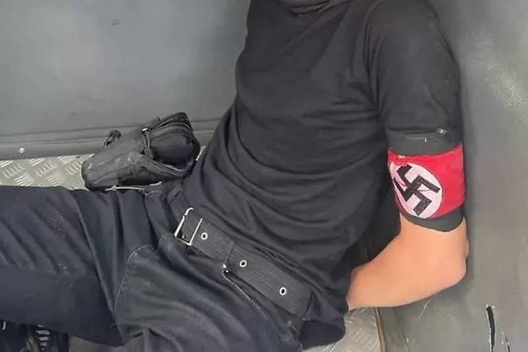 Imagem em close mostra uma pessoa sentada no chão. Ela usa roupa preta e no braço há um bracelete com a suástica nazista