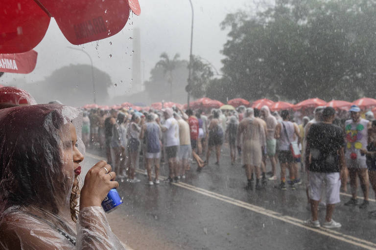Mulher segura lata de bebida. Ela veste capa de chuva. Ao fundo, dezenas de pessoas estão sob forte chuva