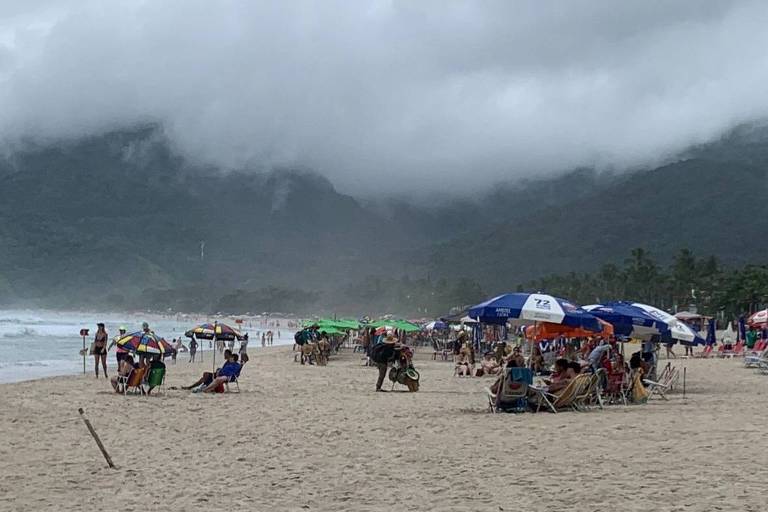 Turistas sob guarda-sol em Maresias. ao fundo, nuvem carregada de chuva encobre parte de morro