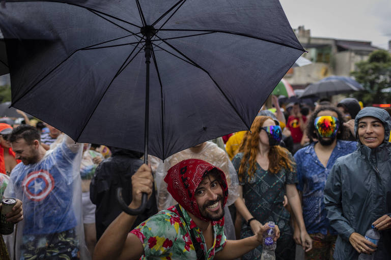folião segurando um guarda chuva preto e bandana na cabeça sorri para a foto em meio a multidão.