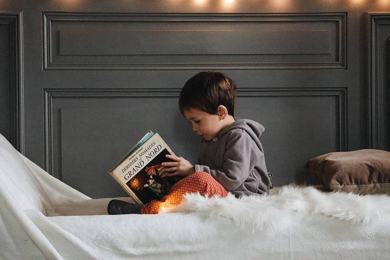 Menino pequeno lendo livro numa cama