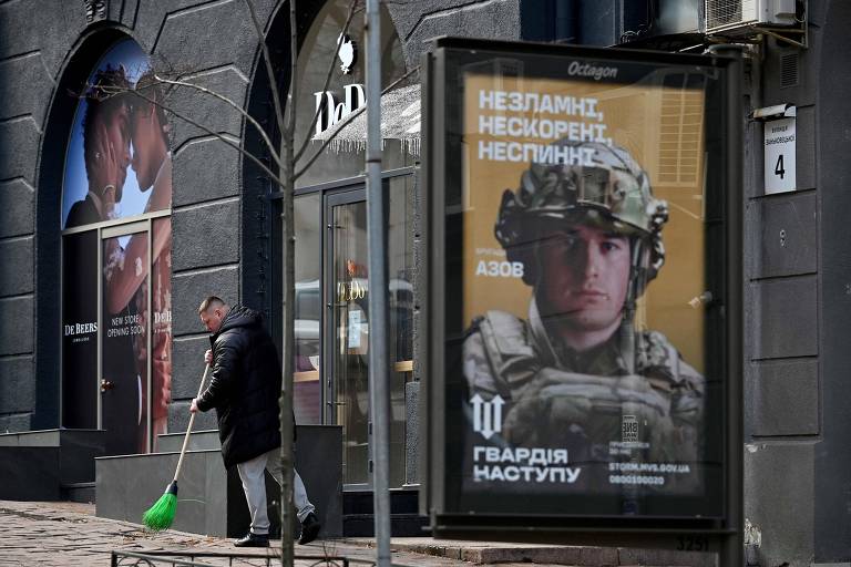 Homem varre calçada na capital Kiev perto de imagem de propaganda com soldado ucraniano e o slogan 'inquebrável, inconquistável, imparável'