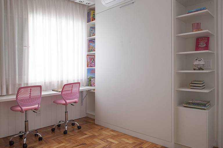 Mesa de estudo encostada na parede com duas cadeiras. No lado direito, um móvel com prateleiras nas laterais e um painel branco
