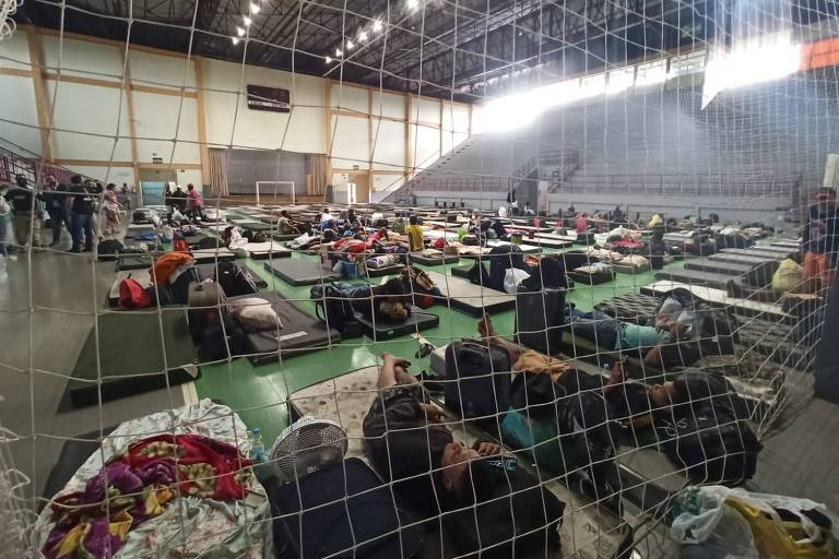 Imagem mostra diversos colchonetes enfileirados sobre quadra de ginásio. Há roupas e algumas pessoas não identificadas dormindo sobre alguns deles.