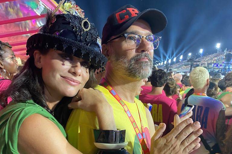 Alinne Moraes, de chapeu com lantejoulas pretas e olhos delineados, aparece abraçando o marido, Mauro Lima, pelas costas; ele usa um boné vermelho e ambos estão no ambiente de um camarote de Carnaval