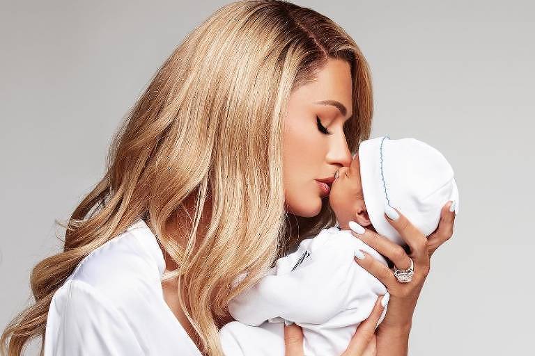 Em foto colorida, mulher de vestido branco e cabelos loiros beija um recém-nascido