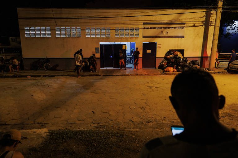 foto mostra escola à noite, é possível ver luz dentro do prédio e pessoas transitando pela entrada. em primeiro plano à direita, uma silhueta de homem que usa o celular 