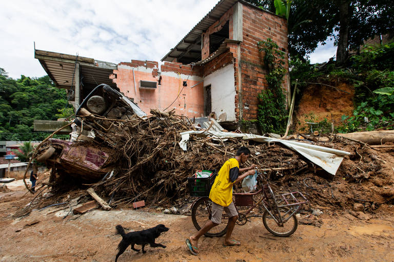 Rapaz empurra bicicleta. Um cachorro preto o segue. Ao fundo, uma pilha de dois metros de lama e entulho e uma casa parcialmente destruída