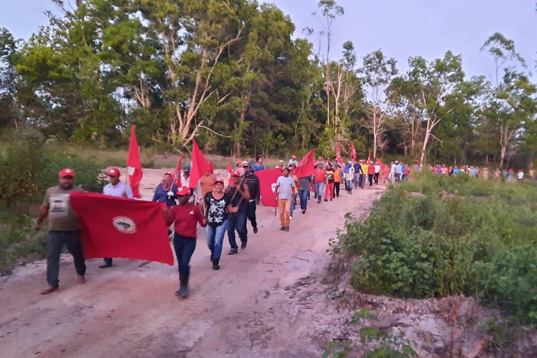 Integrantes do MST caminham em estrada de barro em área rural. Os integrantes seguram bandeiras vermelhas com símbolo do movimento. Às margens, árvores e plantação compõem o espaço.
