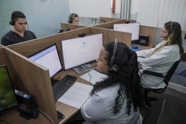 Quatro pessoas, sendo três mulheres de kaleco e um rapaz de blusa preta, todos usando fone em frebte a uma tela de computador, simulam atendimento virtual 