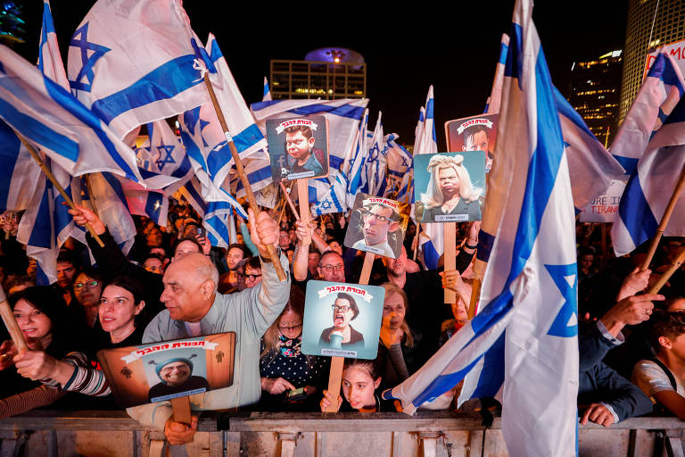 Pessoas se aglutinam em frente a uma mureta segurando cartazes e bandeiras de Israel, durante manifestação 