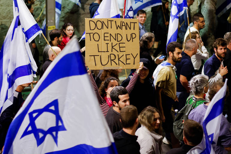 Uma multidão protesta em Tel Aviv carregando bandeiras de Israel (azul e branca) e um cartaz com a mensagem "não queremos terminar como a Turquia"