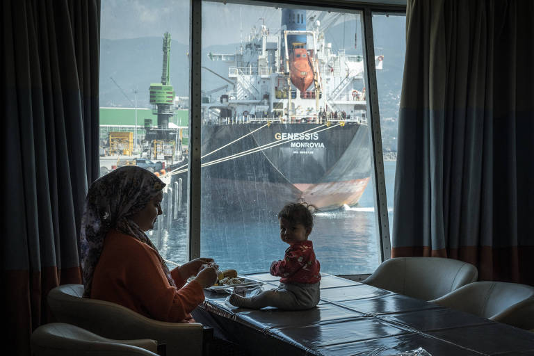 Mulher sentada em cadeira com criança sentada em cima da mesa. Ao fundo da imagem, pela janela, é possível observar outro navio.