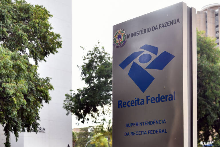 A imagem mostra um edifício com uma placa da Receita Federal do Brasil. A placa é de metal e apresenta o logotipo da Receita Federal, que consiste em um símbolo azul e o texto 'Receita Federal' em letras grandes. Ao fundo, há uma árvore verde e uma parede branca do edifício.