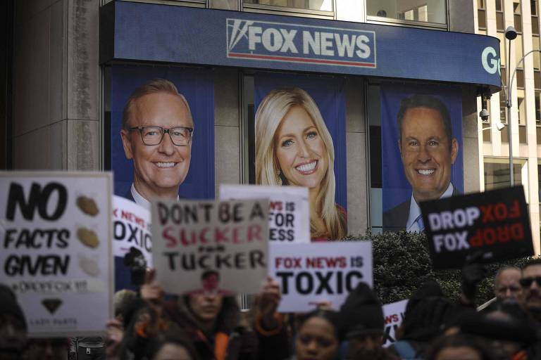 Um grande anúncio de TV na cor azul mostra rostos de apresentadores de programas televisivos; em frente a ele, pessoas protestam com cartazes 