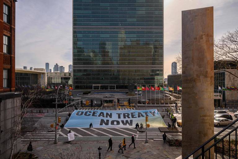 Em frente ao prédio da ONU, de fachada espelhada, manifestantes estendem uma faixa azul clara escrito "Ocean treaty now!"