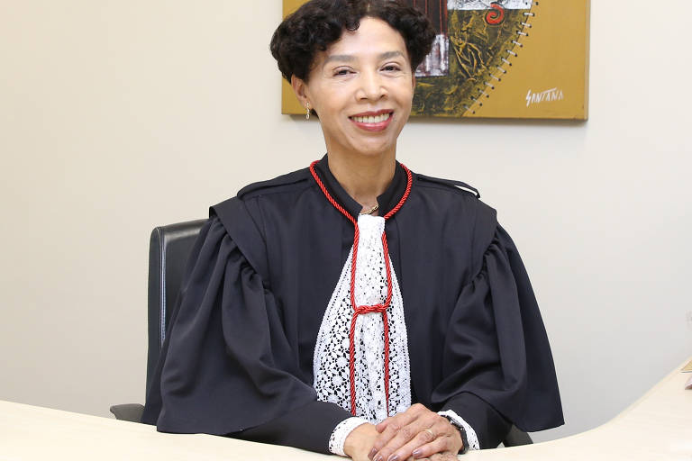 Imagem em primeiro plano mostra uma mulher negra posando para foto. Ela está sentada, com as mãos, uma sobre a outra, apoiadas em uma mesa. Ela usa roupa de juíza na cor preta com detalhes em branco e vermelho.