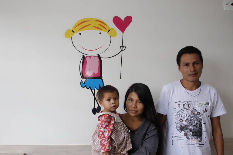 criança, mulher e homem yanomami com desenho colorido de uma criança ao fundo em parede branca