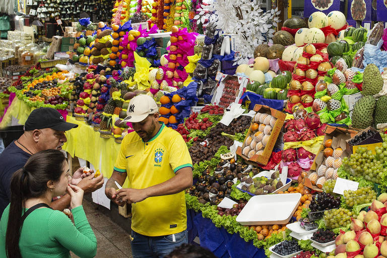 Homem com camiseta da seleção brasileira de futebol corta frutas e oferece a casal, diante de banca com diversas frutas