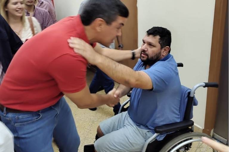 Homem de camiseta vermelha abaixa para se cumprimentar outro homem, de camiseta azul e bermuda jeans, sentado em um cadeira de rodas