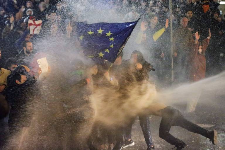 a foto, tirada à noite, mostra manifestantes segurando uma bandeira da União Europeia, formada por um tecido azul com estrelas amarelas dispostas em círculo, sendo dispersados com jatos de água 