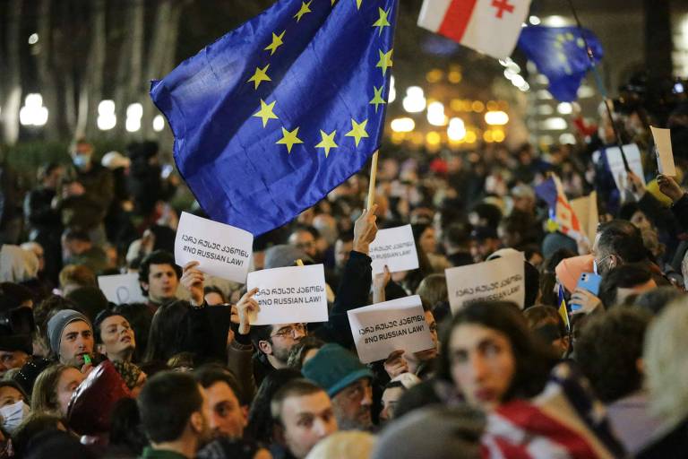 a foto, tirada à noite, mostra uma multidão erguendo cartazes e balançando uma bandeira da União Europeia, pano azul com estrelas amarelas dispostas em círculo