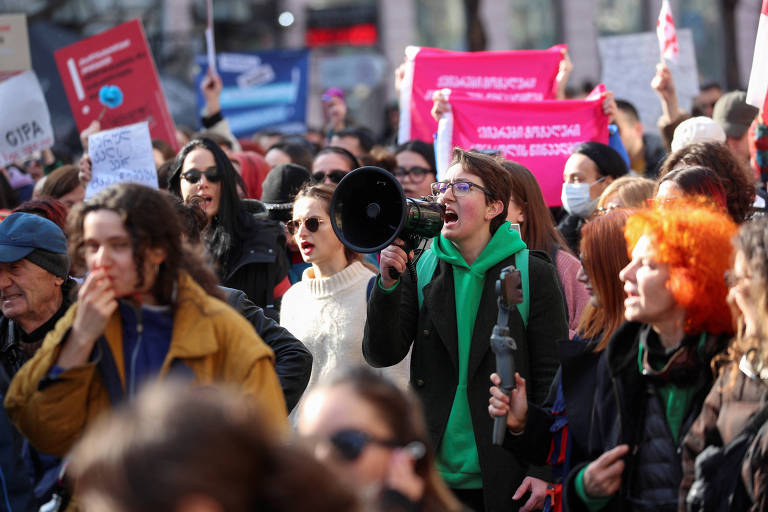 manifestantes, em sua maioria mulheres, são fotografadas enquanto marcham em via pública carregando cartazes e bandeiras. uma delas, ao centro, fala ao megafone