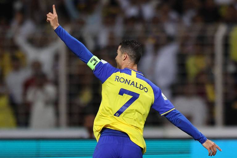 Com o braço esquerdo erguido e o dedo indicador apontado para o alto, Cristiano Ronaldo comemora gol pelo Al Nassr, contra o Al Wehda, no Campeonato Saudita, em partida no estádio Rei Abdulaziz, em Meca; o jogador português usa camisa da cor amarela com o número 7 às costas na cor azul