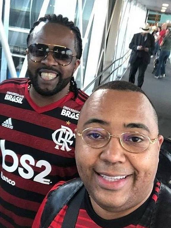 Em foto colorida, dois homens vestidos com camisas do Flamengo posam na entrada de uma estádio de futebol