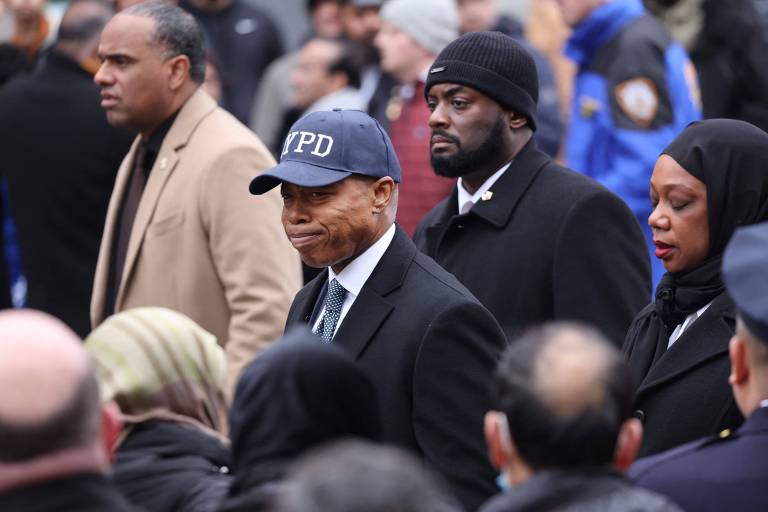 no centro da fotografia, em meio a uma multidão, está Eric Adams, homem negro retinto que usa terno e um boné azul com a sigla NYPD (Departamento de Polícia de Nova York)