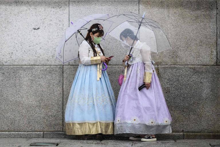Mulheres com roupas tradicionais coreanas, coloridas e com saias até o pé, seguram guarda-chuva e olham para o chão