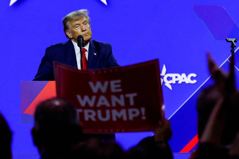 Trump, homem branco de pele alaranjada e cabelos loiros, discursa em cima de um púlpito iluminado. uma pessoa na plateia à sua frente ergue um cartaz onde se lê "we want trump" ("nós queremos trump")