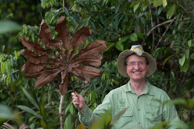 Homem idoso posa sorrindo enquanto segura uma folha grande e seca com a mão direita