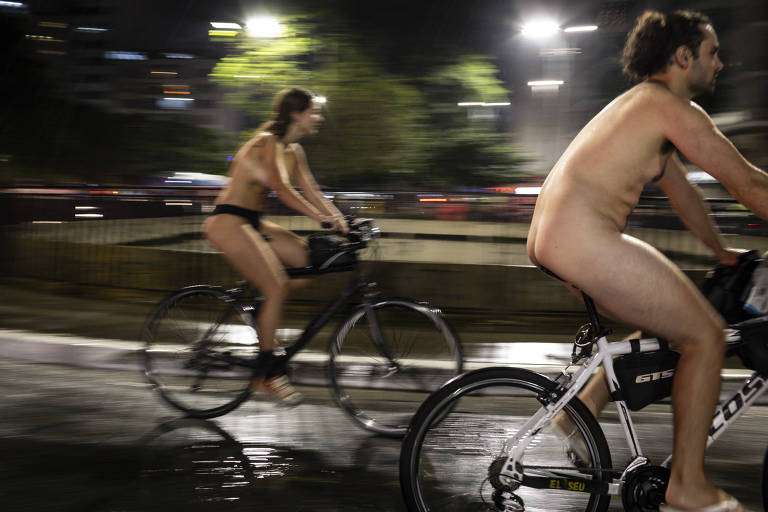 Mulher e homem nus, cada um numa bicicleta, pedalam à noite em frente a vão na praça do Ciclista