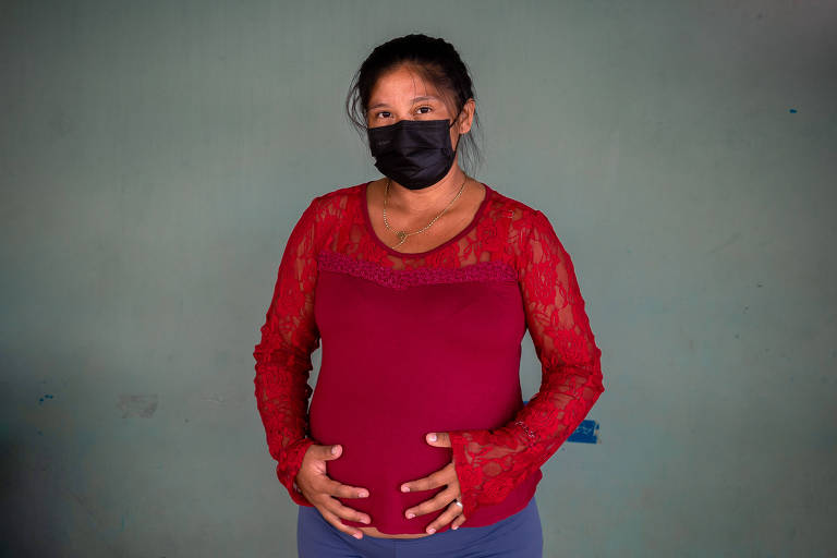 Indígena gestante, vestindo blusa vermelha e calça azul, segura a barriga, enquanto espera por atendimento de pré-natal em posto de saúde em Boa Vista, capital de Roraima