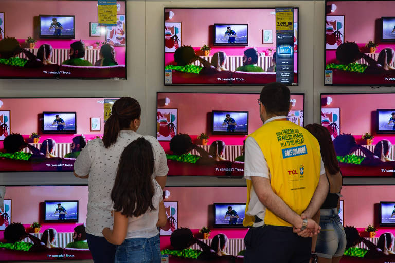 Vendedor, consumidora e criança olha para parede cheia de TVs em exposição, ligadas