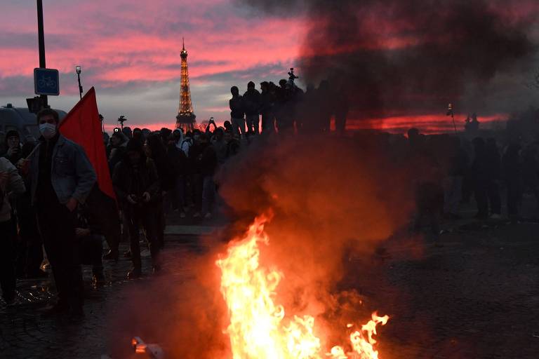 ao entardecer, manifestantes ateiam fogo em barreira. ao fundo, a torre eiffel aparece junto ao pôr-do-sol