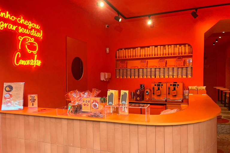 A foto mostra um balcão com produtos de chocolate e um salão pintado de laranja com um neon amarelo na parede