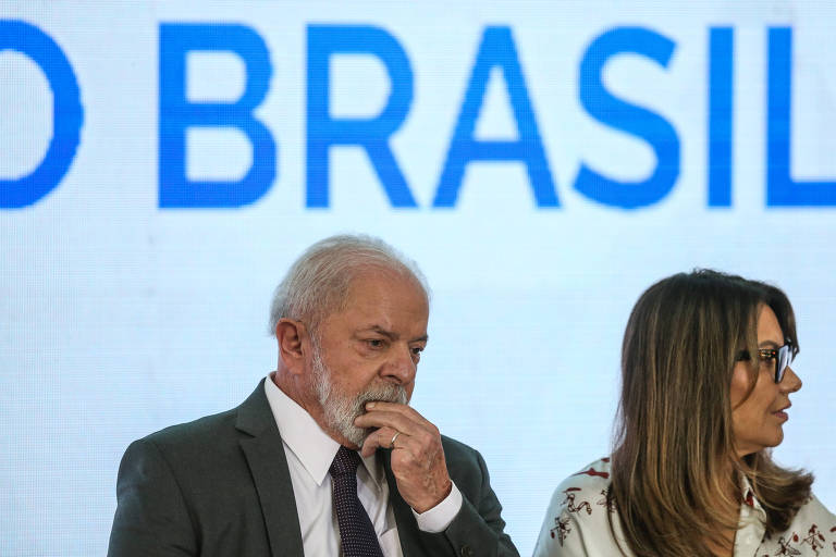 O presidente Lula olha para baixo e coloca a mão no queixo; Janja, sentada perto dele, olha para o lado; ao fundo, aparece o texto "o Brasil"