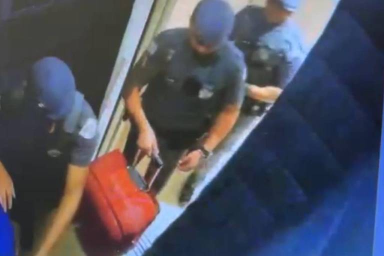 Policiais militares entram no elevador com uma mala grande vermelha