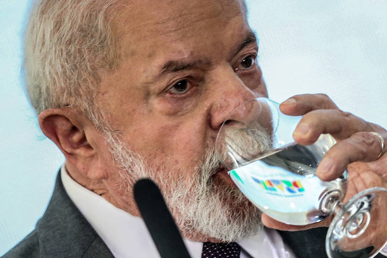 Lula, um homem branco de cabelos grisalhos, bebe água em uma taça. Ele veste terno chumbo, camisa branca e gravata escura.