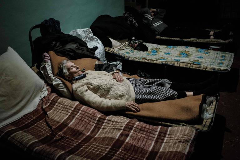 uma mulher idosa aparece dormindo numa cama improvisada dentro de um bunker, sobre cobertores. outras três camas foram dispostas ao seu lado