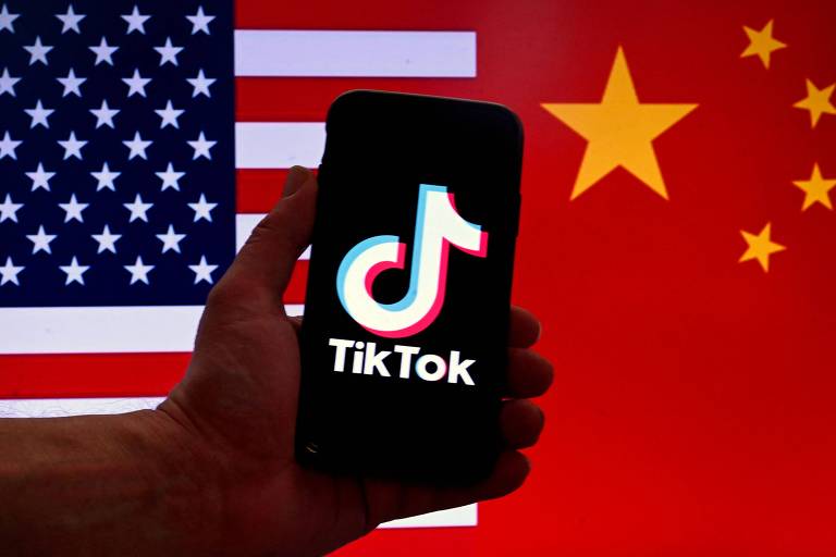 Mão segura celular, cuja tela mostra o aplicativo do TikTok. Ao fundo, estão uma bandeira dos Estados Unidos e outra da China, repartindo o quadro pela metade.