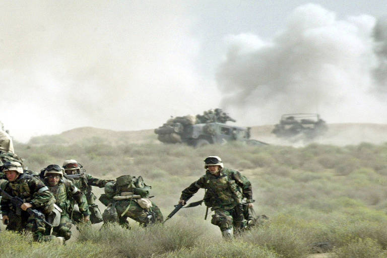 num campo de guerra, soldados correm em meio à mata rasteira. ao fundo, é possível ver tanques e fumaça