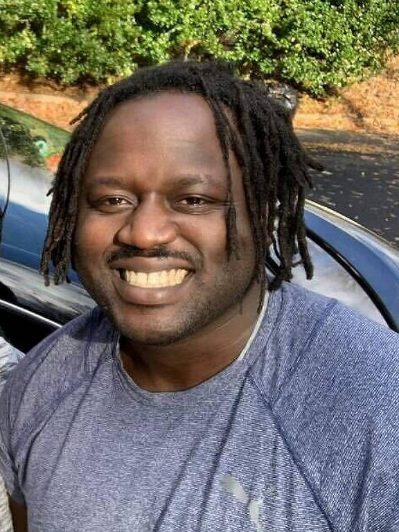a foto mostra Otieno, um homem negro retinto que sorri. ele usa o cabelo caído em pequenos dreads e está com uma camiseta cinza