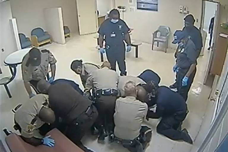 na imagem, registro de uma câmera, estão diversos policiais e funcionários sobre o corpo de um homem negro, escondido abaixo das pessoas